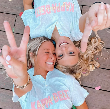 Two women lying down wearing matching grey Kappa Delta big little reveal button-up shirts