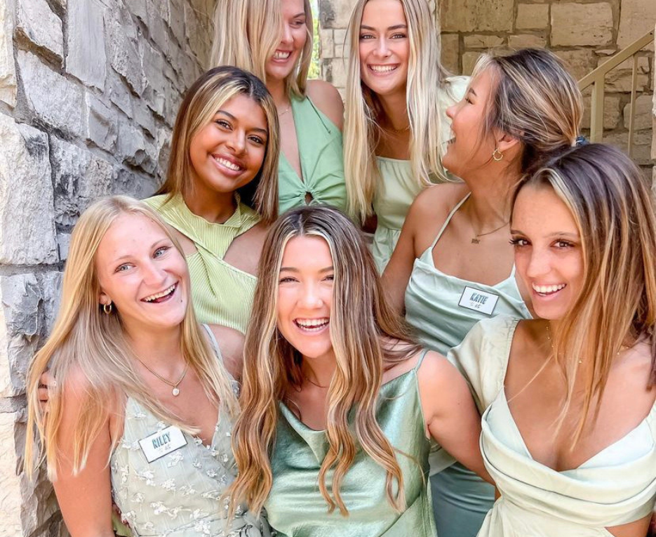 Seven women stand together in sage colored dresses