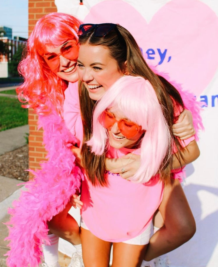 Three girls stand together hugging in pink shirts and wigs.