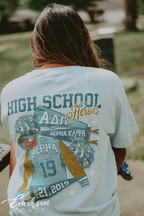 Girl wears a light blue shirt featuring a letterman jacket, pom poms, and a bullhorn that reads High School Heroes.